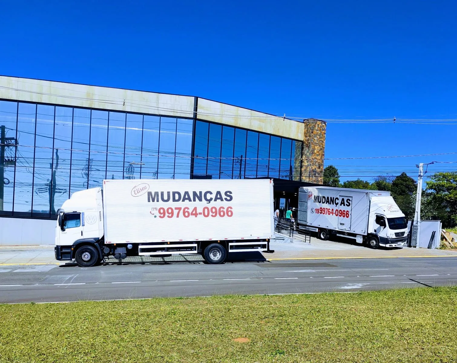 Dois caminhões baú da Eliseu Mudanças estacionados em frente a um prédio comercial de fachada de vidro